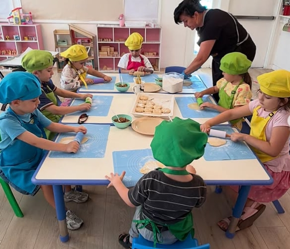 Children wearing colorful chef hats and aprons rolling dough during a Little Chef cooking class